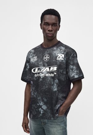 Мъж позира с черно-сива тениска Off-White с десен и бели лога, както и със сини дънки, на изчистен светъл фон.