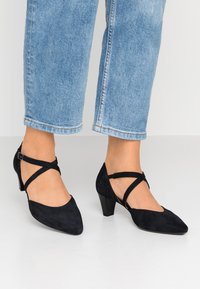 Scarpe nere in suede con tacco alto e design a punta, caratterizzate da cinturini incrociati e un piccolo tacco a blocco. Indossate con jeans a vita alta di colore blu chiaro.