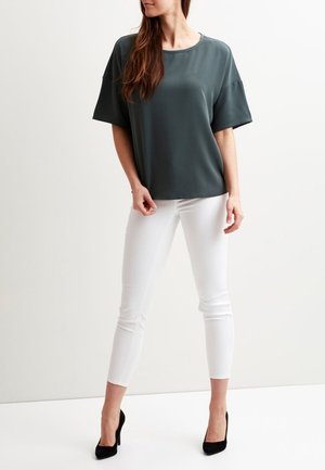 Blouse verte foncée à manches courtes et pantalon blanc ajusté, assortis à des escarpins noirs. La blouse présente un col rond et une texture lisse.