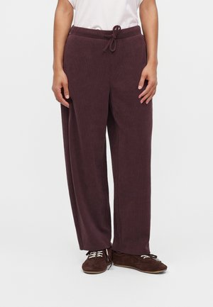 Personne portant un pantalon de survêtement ample violet foncé avec une taille à cordon, un haut blanc et des chaussures marron foncé à lacets, debout devant un fond uni.