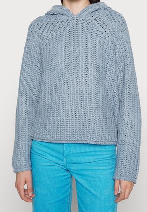 Personne portant un pull à capuche en grosse maille bleu clair et un pantalon en velours côtelé turquoise vif, debout les bras détendus le long du corps.