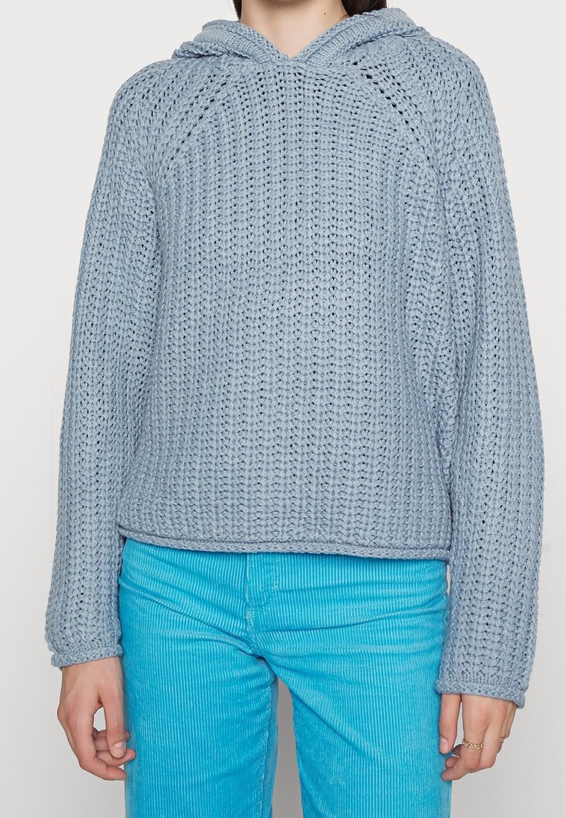 Personne portant un pull à capuche en grosse maille bleu clair et un pantalon en velours côtelé turquoise vif, debout les bras détendus le long du corps.