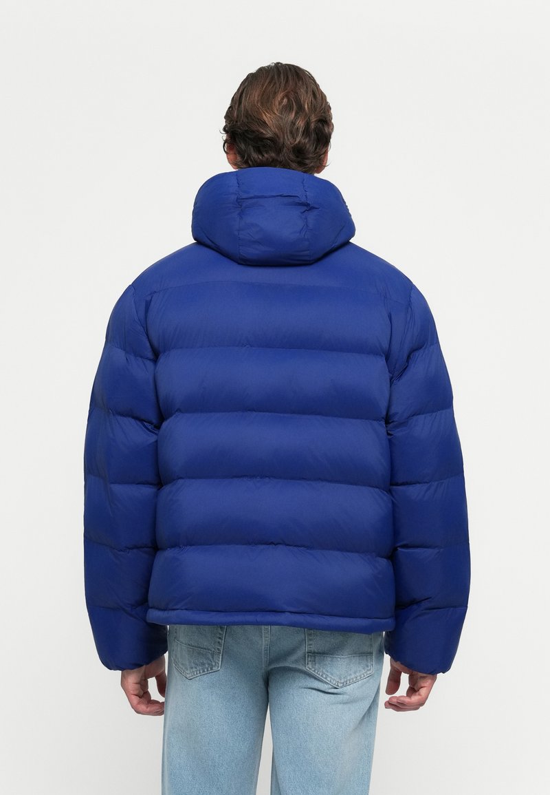 Kék puffernadrág, magas gallérral és vízszintes steppelt mintával. Simább anyagból készült, kapucnival és széles ujjakkal.