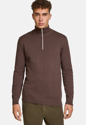 Uomo con maglione marrone testurizzato con zip, polsini e orlo a coste, abbinato a pantaloni verde oliva, in piedi su sfondo semplice.