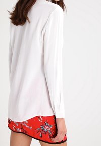 Blusa bianca a maniche lunghe con una texture liscia, caratterizzata da un orlo curvo. Abbinata a una gonna rossa a fantasia floreale con bordo nero.