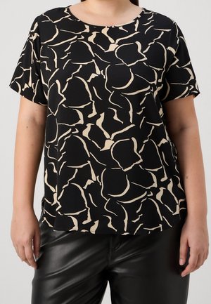 Vrouw draagt een zwart topje met korte mouwen en een abstract beige patroon, gecombineerd met zwarte leren broek, tegen een effen achtergrond.