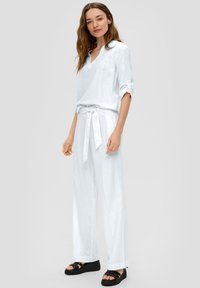 Haut en lin blanc avec un décolleté en V et des manches retroussées, associé à un pantalon large assorti avec une taille à nouer. Chaussures noires visibles.