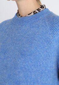 Niebieski dzianinowy sweter o prążkowanej fakturze, z okrągłym dekoltem. Z pod spodu wystaje koszula w leoparda z kołnierzykiem.