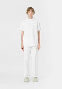 Camiseta blanca de gran tamaño con logotipo en relieve, combinada con pantalones blancos de corte holgado, que presentan un detalle de cadena. Llevado con zapatos de textura en tonos claros.