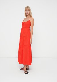 Rode maxi-jurk met dunne bandjes, gevormde lijfje en gelaagde rok. Gemaakt van lichtgewicht stof met een subtiele textuur. Dragen met zwarte slippers.