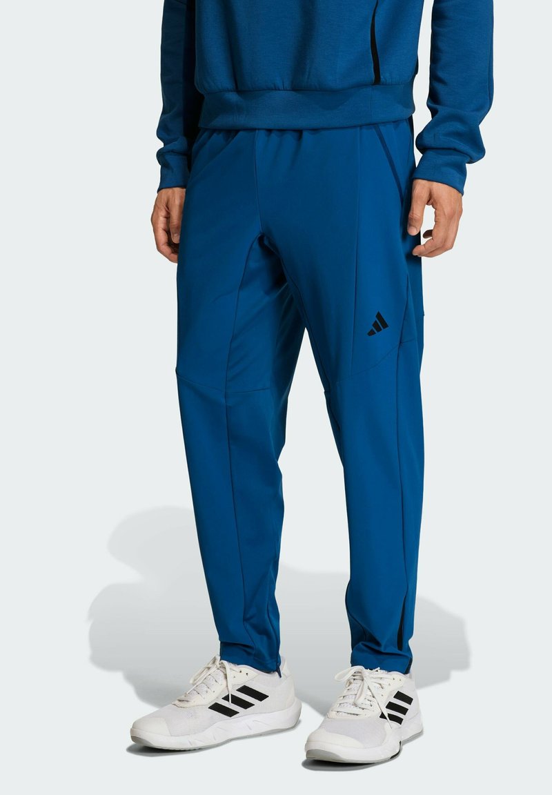 Blaue Sporthose aus dehnbarem Material, mit Seitentaschen und schwarzem Logodetail. Kombiniert mit weißen Turnschuhen.
