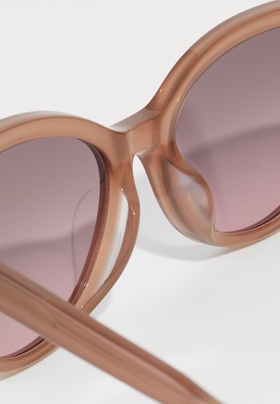 Ochelari de soare supradimensionați cu un cadru translucid de culoare maro deschis și lentile gradient roz, având o finisare netedă și lucioasă și un design curbat.