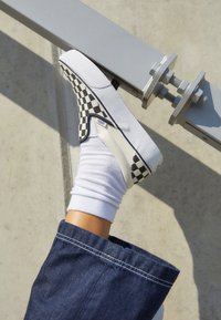 Crno-bijeli karo uzorak slip-on tenisice s glatkom platnenom gornjom, naglašenom bijelom potplatom, nošene s bijelom čarapom i trapericama.