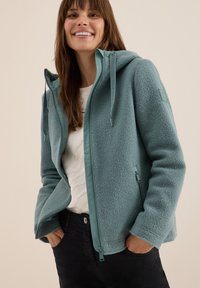 Veste en polaire teal clair avec une fermeture éclair sur le devant, une capuche et deux poches latérales. Elle possède une doublure lisse et un logo sur la manche.