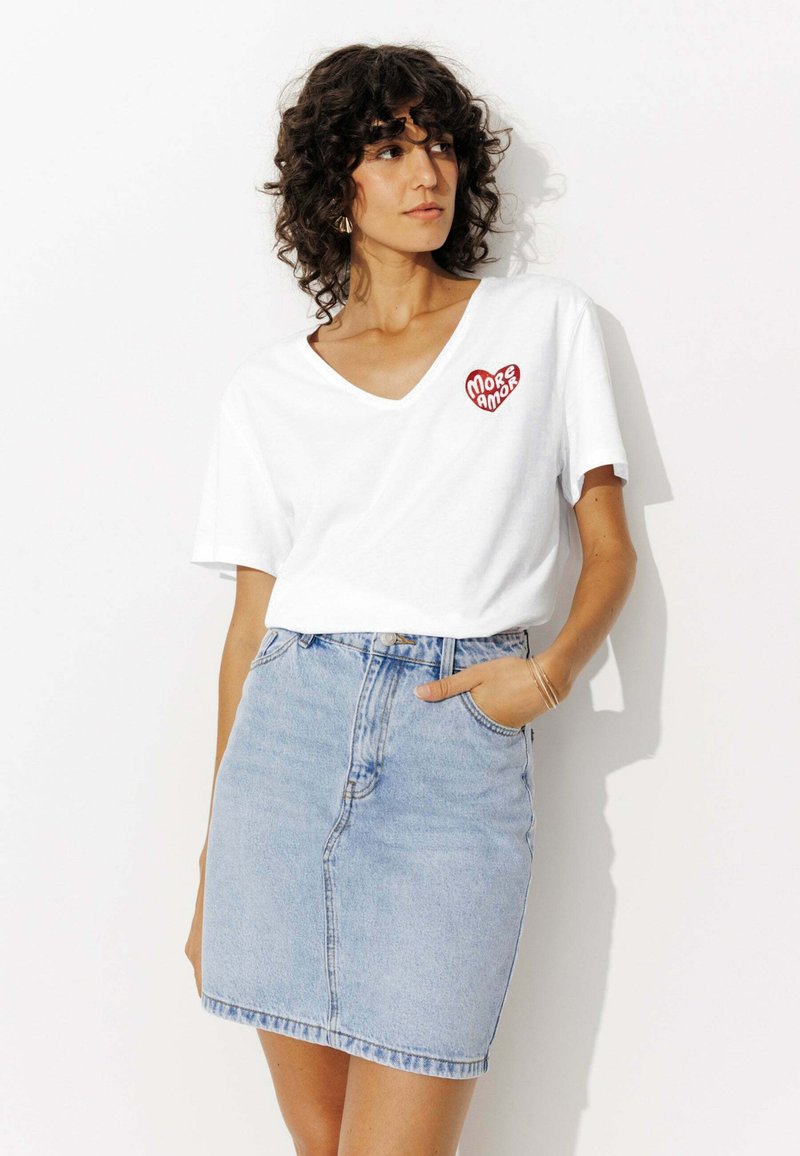 T-shirt en coton blanc avec un col en V et un design brodé en forme de cœur rouge "More Amor", associé à une mini jupe en jean bleu clair.