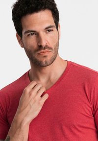 Muž s krátkými kudrnatými tmavými vlasy a vousy, oblečený v červeném tričku s výstřihem do V, dotýká se límce a zamyšleně hledí stranou na bílém pozadí.