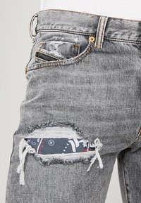 Grå slitna jeans med ett synligt blått och vitt mönstrat tygbit under tåren nära övre låret och en liten bakficka.
