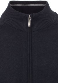 Marinblå zip-up tröja med hög krage, ribbad textur och en silverfärgad dragkedja. Har kontrasterande randig detalj inuti kragen.