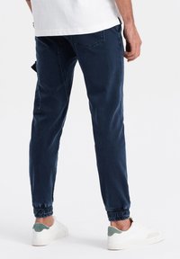 Pantalones jogger azul marino de mezcla de algodón con puños elásticos, que cuentan con un bolsillo trasero y una textura sutil, llevados con zapatillas blancas.