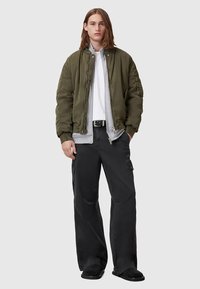 Olivgrüne Bomberjacke mit geripptem Kragen, darunter hervorlugende graue Kapuzenjacke, kombiniert mit schwarzen Cargo-Hosen und schwarzen Slippern.