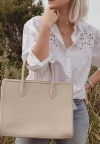 Beige leren tote bag met een gestructureerd ontwerp, verlengde handvaten en een geëmbosserd logo. Draagt door een persoon in een wit shirt en jeans.