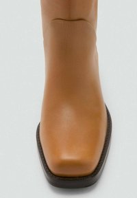 Bottine en cuir tan avec un bout carré, texture lisse et semelle foncée. Les coutures le long des côtés mettent en valeur le design.