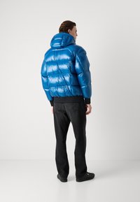 Zils pūkains mētelis ar spīdīgu, quilted dizainu, kura sastāvā ir cepure un ribotas aproces. Apvienots ar melniem džinsiem un tumšām kurpēm.