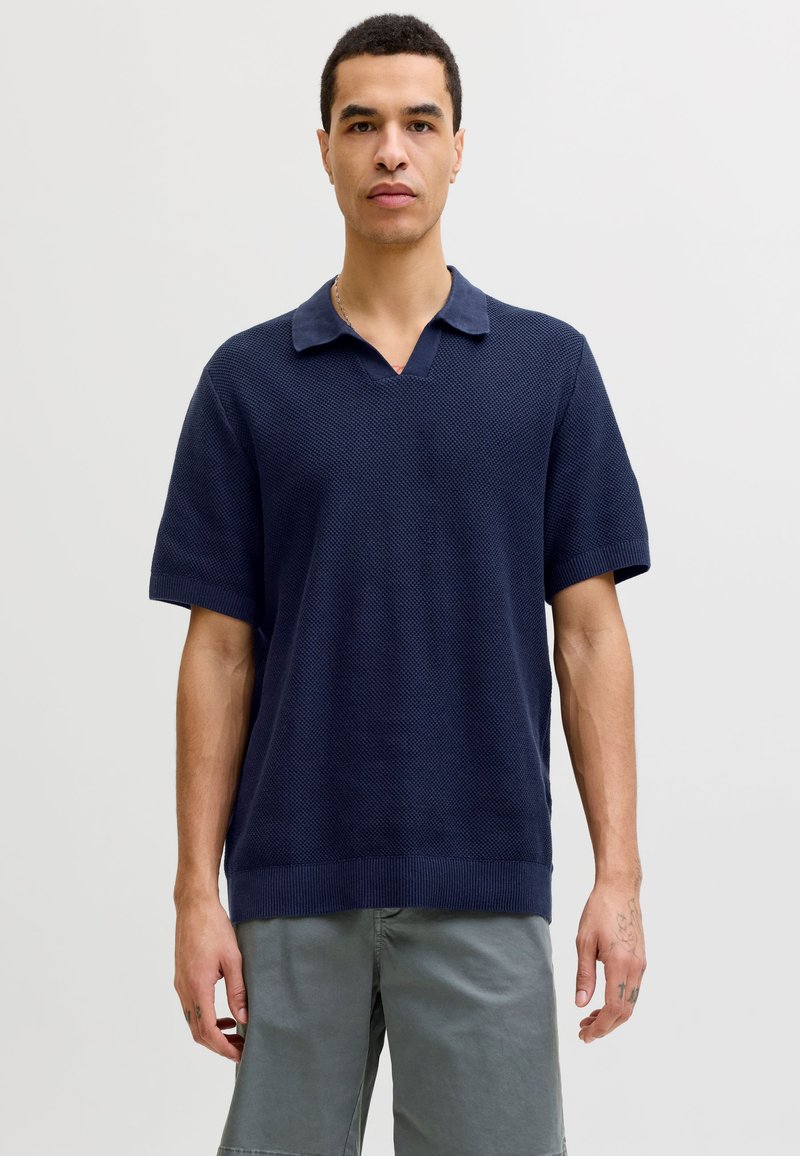 Homme portant un polo en maille bleu marine à manches courtes et un short gris, debout avec une expression neutre sur un fond uni clair.