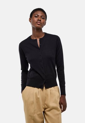 Femme portant un cardigan noir boutonné et un pantalon beige plissé à taille haute, debout les mains dans les poches devant un fond uni.