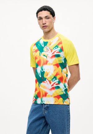 Jonge man met kort krullend haar die een kleurrijk, abstract sportshirt met gele mouwen en blauwe spijkerbroek draagt, staand met handen achter de rug.