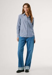 Camicia a maniche lunghe a righe blu e bianche con colletto, abbinata a jeans in denim blu a gamba larga e scarpe nere.