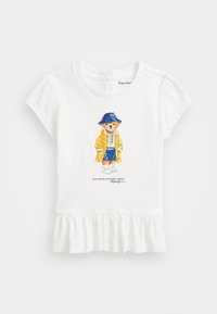 Biała koszulka z krótkim rękawem z falbaną na dole, z nadrukiem misia w żółtym płaszczu przeciwdeszczowym i niebieskiej czapce, w paski koszulce i trampkach.