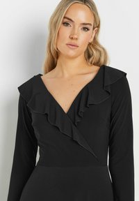 Czarna sukienka z długim rękawem, z głębokim dekoltem w kształcie V i falbankami wzdłuż linii dekoltu, wykonana z gładkiego, miękkiego materiału.