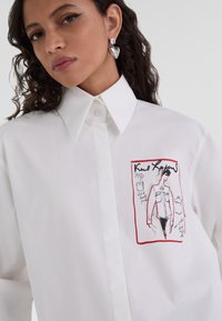 Camicia bianca con bottone e design illustrato, decorata con dettagli rossi. Il tessuto sembra liscio e presenta un colletto strutturato. Orecchini a forma di cuore in argento.