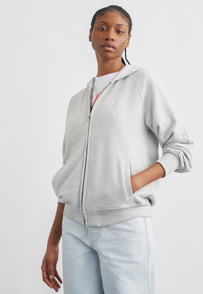 Mladá osoba s copánkami, ktorá má na sebe svetlosivú mikinu na zips a svetlomodré rifle, stojí s jednou rukou v vrecku.