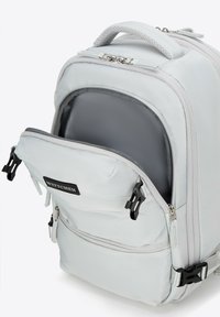 Sac à dos gris clair en tissu résistant ; dispose d'une poche avant avec des doubles zips, d'une poignée rembourrée et d'un écusson de marque noir.