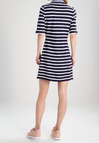 Mujer vestida con un vestido de rayas azul marino y blanco de manga corta y zapatillas rosas, de pie con la espalda a la cámara sobre un fondo blanco.
