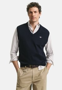 Marineblauwe wollen vest met een V-hals, gedragen over een wit en rood gestreept overhemd met kraag, gecombineerd met kaki broek en een bruine riem.