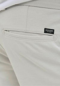 Světle šedé kalhoty s kapsou na zadní straně se štěrbinovým designem a černou etikou s nápisem "JACK & JONES EST. 1990" našitou na ní.