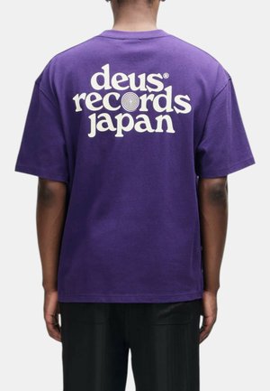 Persona che indossa una maglietta viola con testo bianco "deus records japan" e un motivo a spirale sulla schiena, rivolta con la schiena alla fotocamera.