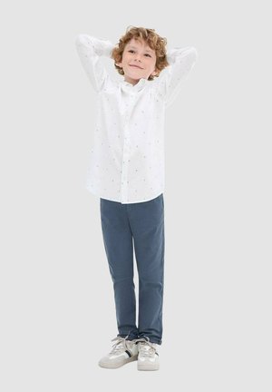 Camicia bianca con piccoli pois blu, abbinata a pantaloni blu navy e scarpe da ginnastica chiare. Il ragazzo è in piedi con le braccia dietro la testa.