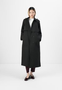 Cappotto lungo nero con vestibilità ampia, caratterizzato da un ampio colletto, maniche lunghe e tasche laterali, indossato sopra un top bianco e pantaloni bordeaux.
