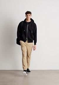 Zwarte zip-up hoodie, beige cargobroek met zakken en zwarte sneakers. Model staat tegen een neutrale achtergrond en toont casual kleding.