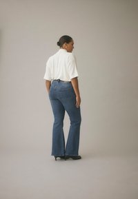 Traperice visokog struka plave boje s teksturiranim završetkom i širokim nogavicama, uparene s bijelom širokom majicom. Model nosi crne cipele s potpeticom.