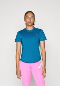 Egy nő áll egy fehér háttér előtt, kék Nike pólóban és élénk rózsaszín leggingsben, amelyen a Nike logó látható.