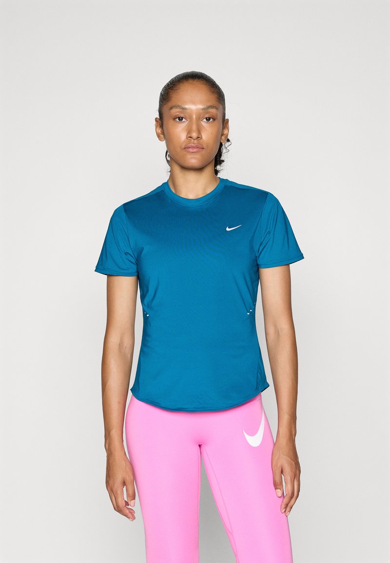 Egy nő áll egy fehér háttér előtt, kék Nike pólóban és élénk rózsaszín leggingsben, amelyen a Nike logó látható.