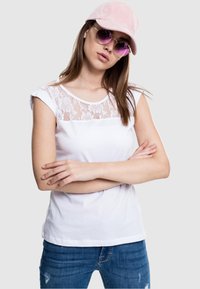 Camiseta blanca con panel de encaje en los hombros, de mangas cortas, combinada con vaqueros azules desgastados. El modelo lleva como accesorios una gorra rosa y gafas de sol.