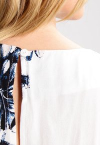 Top bianco con chiusura a bottone sul retro, caratterizzato da un motivo floreale a contrasto in nero e blu su un lato. Tessuto morbido e testurizzato.