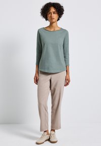 Hellgrünes Langarmshirt mit rundem Ausschnitt, kombiniert mit beigen maßgeschneiderten Hosen und hellen Sneakers, auf einem neutralen Hintergrund präsentiert.