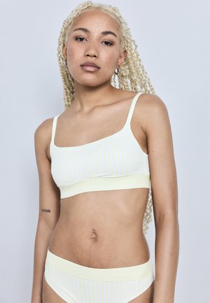 Νεαρή γυναίκα με μακριά ξανθά πλεξούδια που φοράει ανοιχτό κίτρινο ριγέ μπικίνι, στέκεται μπροστά σε απλό ανοιχτό γκρι φόντο.